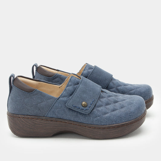 A pair of women's slip-on shoes in a dusty blue quilted fabric with a brown wedge sole and a small gold-tone snap detail on the strap, shown against a light gray background.