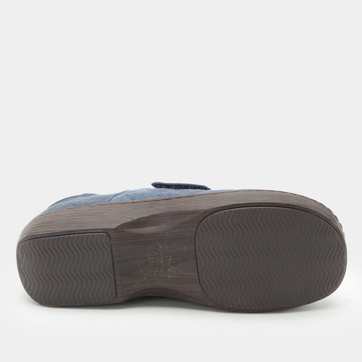 A close-up bottom view of a slip-on shoe or clog, showing a dark brown textured sole with a wavy tread pattern and a light blue fabric upper against a light gray background.
