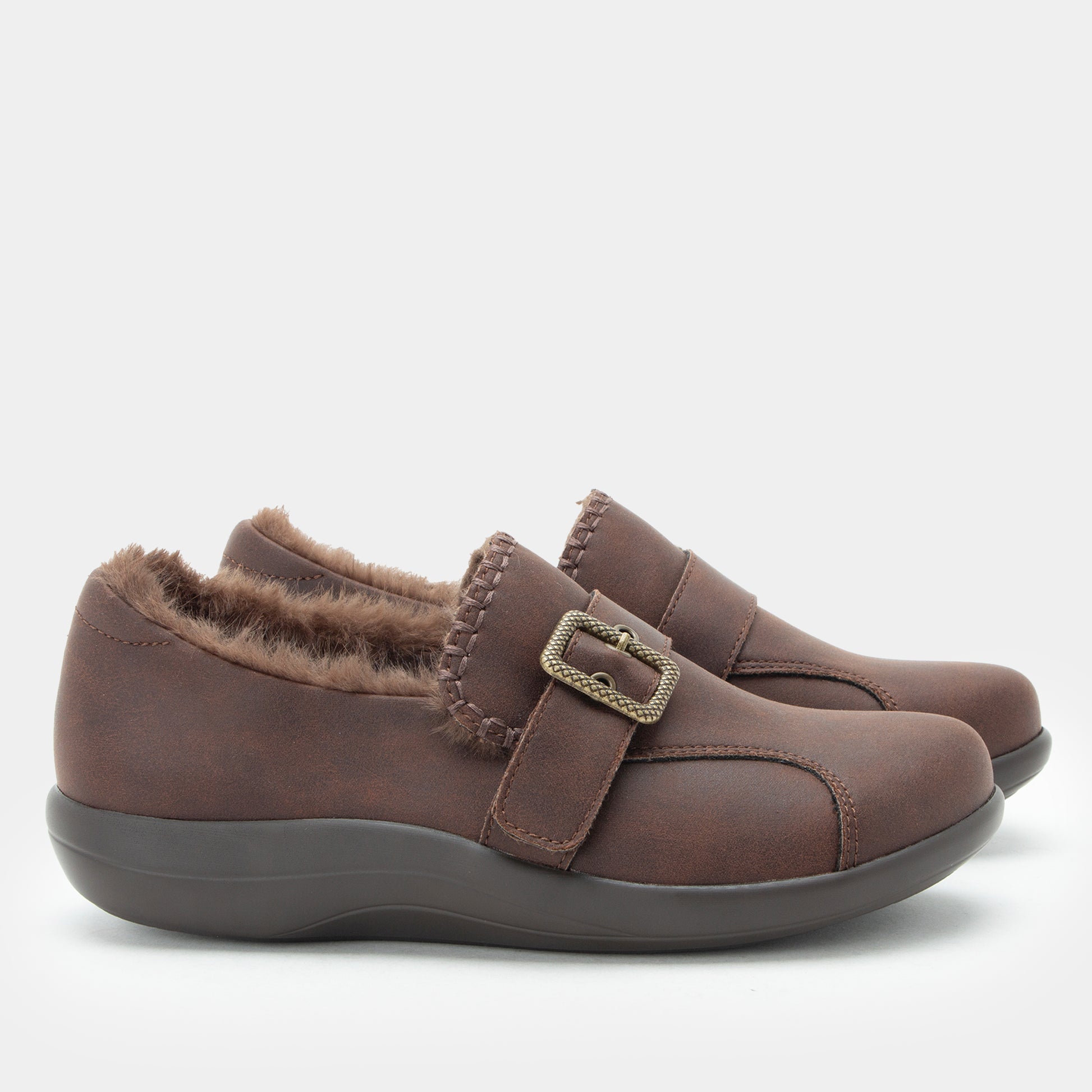 A pair of women's slip-on shoes in dark brown faux leather with a fuzzy brown shearling-style lining and a decorative metal buckle strap, shown against a light gray background.