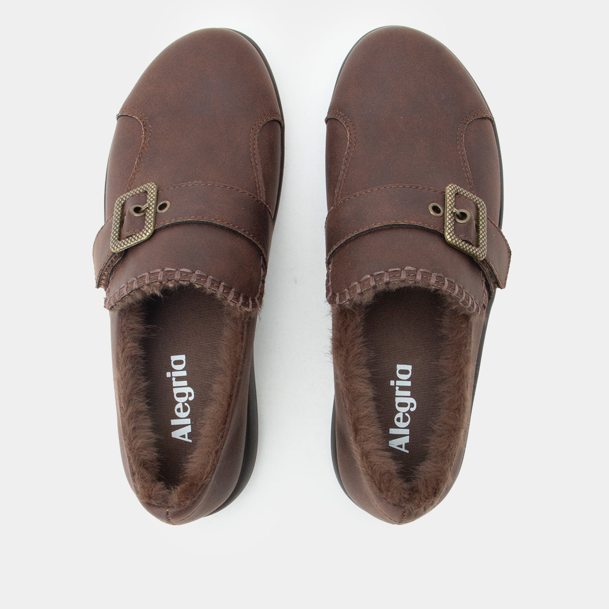 A pair of dark brown slip-on clogs with faux fur lining and decorative buckle straps, shown from a top-down view on a light gray background. The brand name 'Alegria' is printed on the insole.