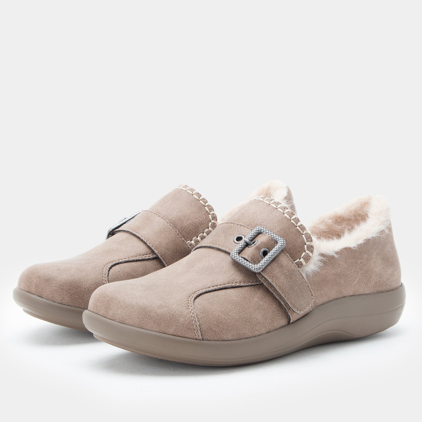 A pair of taupe suede slip-on shoes with a plush white shearling lining and a decorative silver buckle strap, shown on a light gray background.