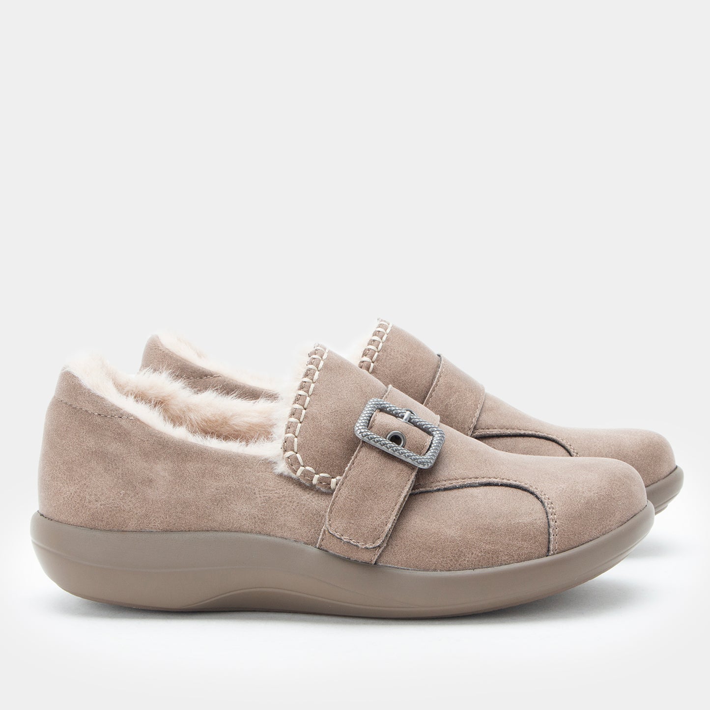 A pair of taupe suede slip-on shoes with a plush cream lining and a decorative silver buckle strap, shown against a light gray background.