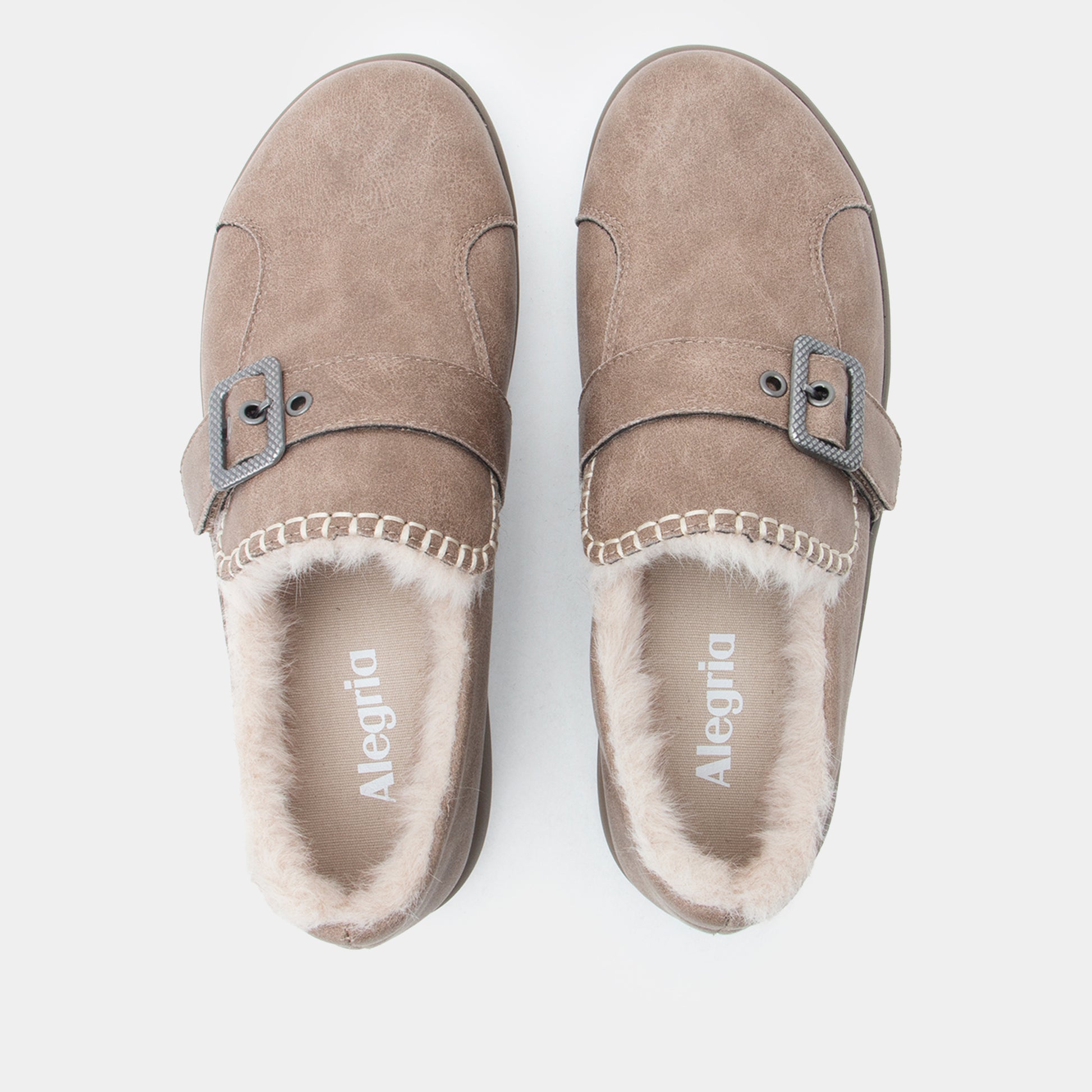 A pair of Alegria taupe suede slip-on mule slippers with faux fur lining and a decorative strap with a buckle, shown from a top-down view on a light gray background.