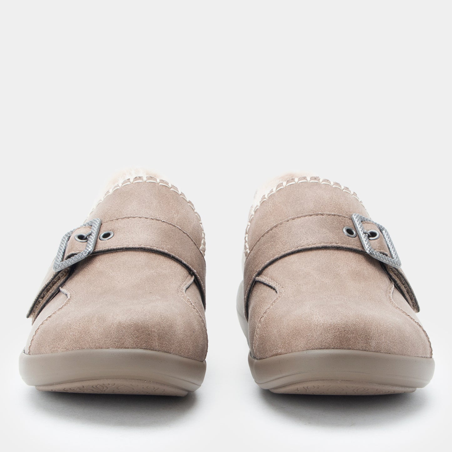 A pair of taupe suede slip-on shoes with double straps and silver buckle details, shown from a front-facing perspective on a light gray background.