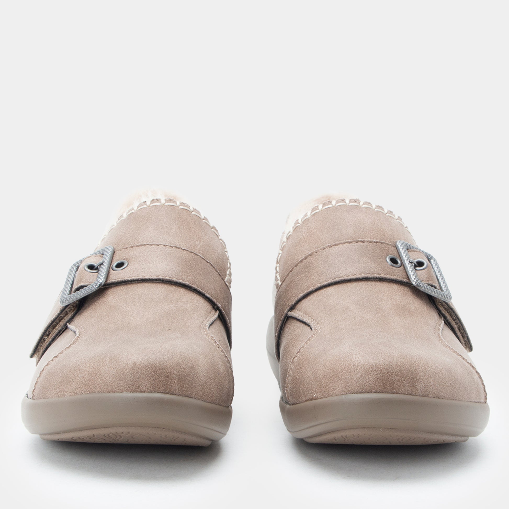 A pair of taupe suede slip-on shoes with double straps and silver buckle details, shown from a front-facing perspective on a light gray background.