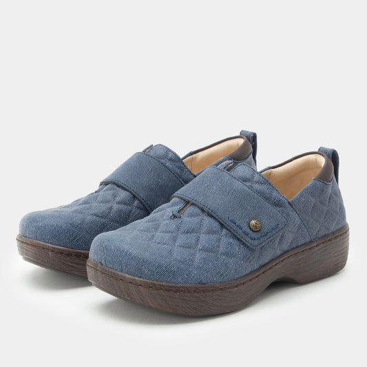 A pair of women's slip-on shoes in a quilted blue fabric with dark brown soles and a small gold button detail on the strap, shown against a light gray background.