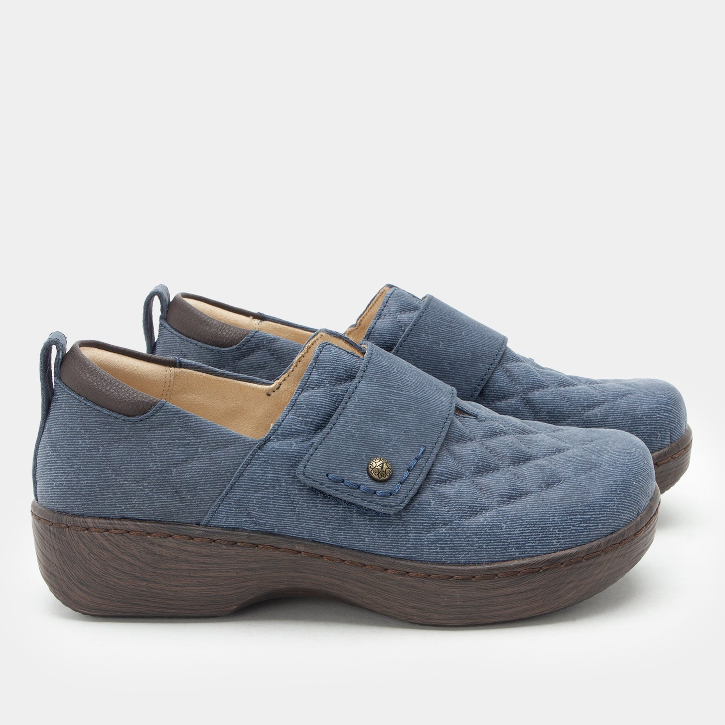 A pair of women's slip-on shoes in a dusty blue quilted fabric with a brown wedge sole and a small gold-tone snap detail on the strap, shown against a light gray background.