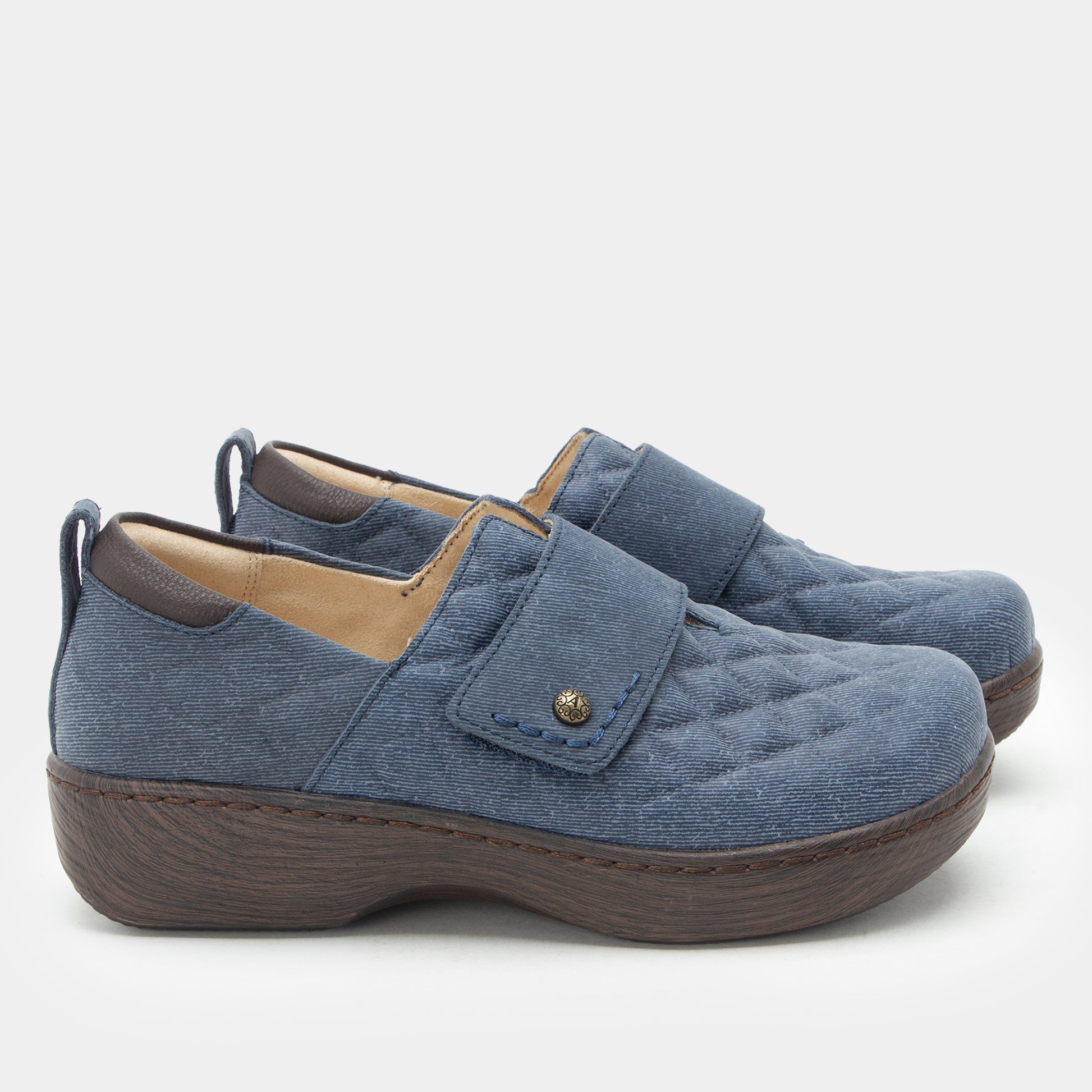 A pair of women's slip-on shoes in a dusty blue quilted fabric with a brown wedge sole and a small gold-tone snap detail on the strap, shown against a light gray background.