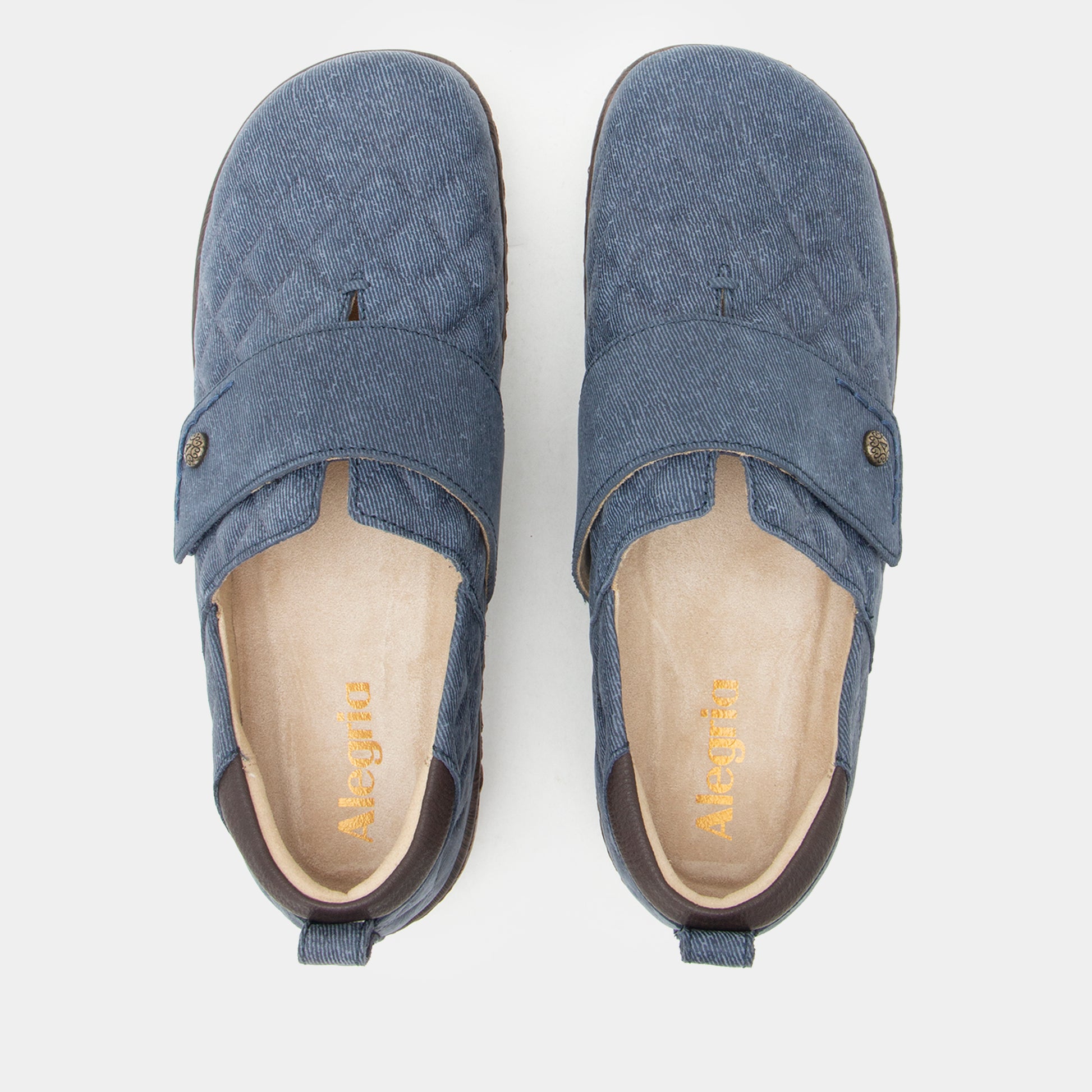 A pair of blue quilted slip-on shoes with a wide strap and pull tabs, shown from a top-down view on a light gray background. The brand name 'Meggi' is visible in gold on the beige insoles.