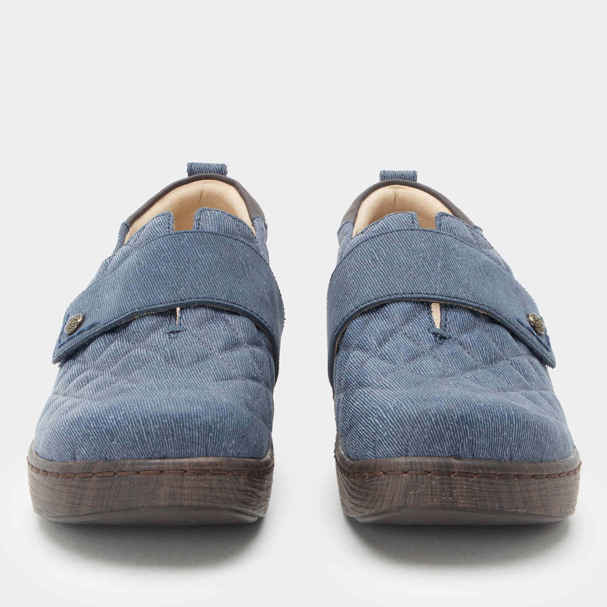 A pair of slip-on quilted shoes in a dusty blue fabric, featuring a hook-and-loop strap closure with small gold-tone metal accents and dark brown soles, shown against a light gray background.