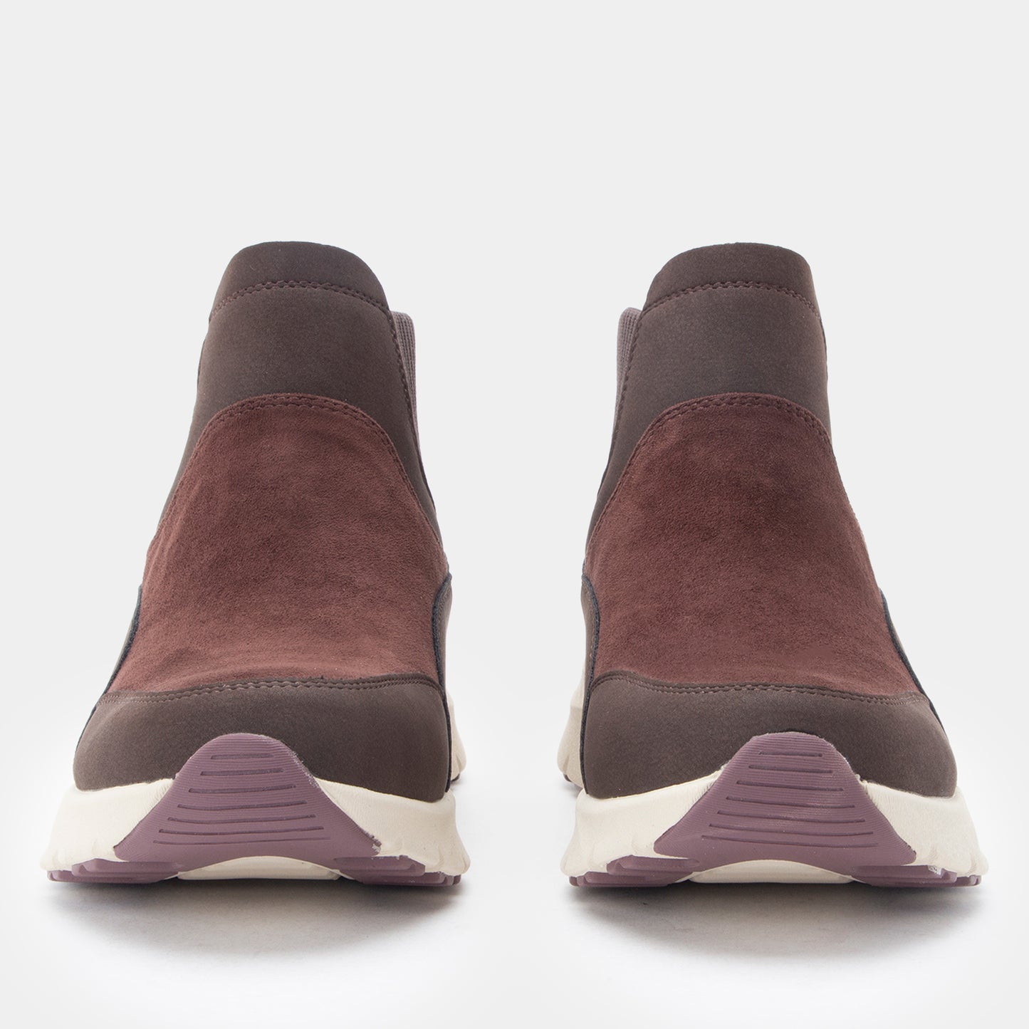 A pair of slip-on ankle boots shown from the front on a light gray background. The boots feature a dark brown suede upper, a darker brown elastic side panel, and a thick white sole with a muted purple textured accent at the bottom.