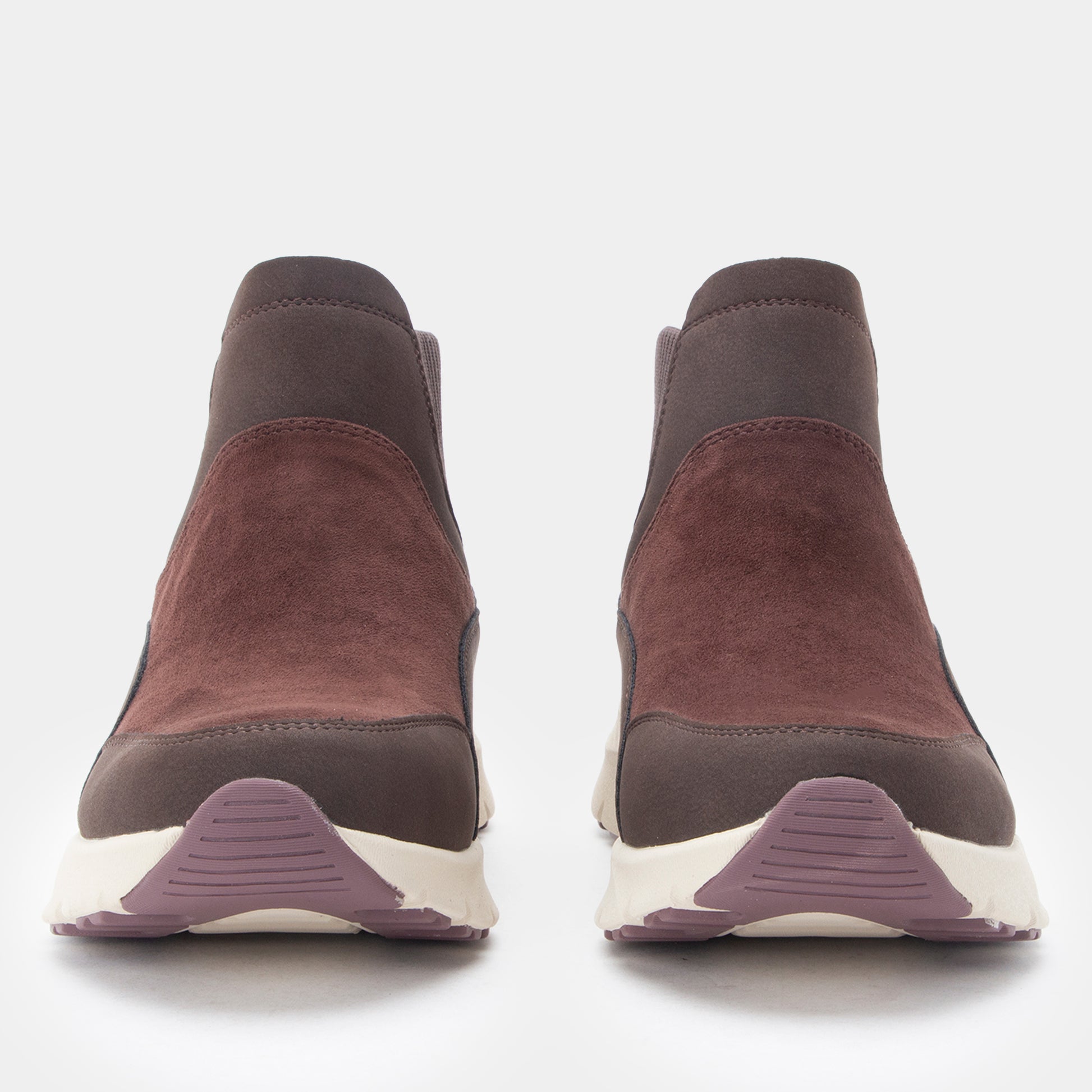 A pair of slip-on ankle boots shown from the front on a light gray background. The boots feature a dark brown suede upper, a darker brown elastic side panel, and a thick white sole with a muted purple textured accent at the bottom.