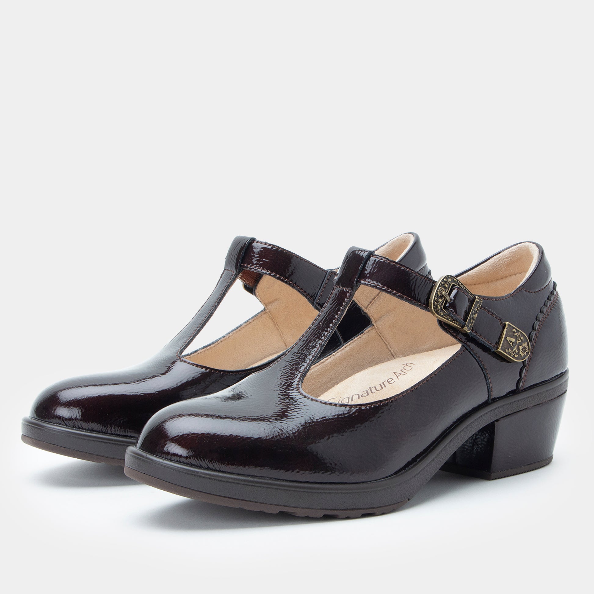 A pair of dark brown patent leather Mary Jane shoes with low block heels and buckle straps, shown on a light gray background.