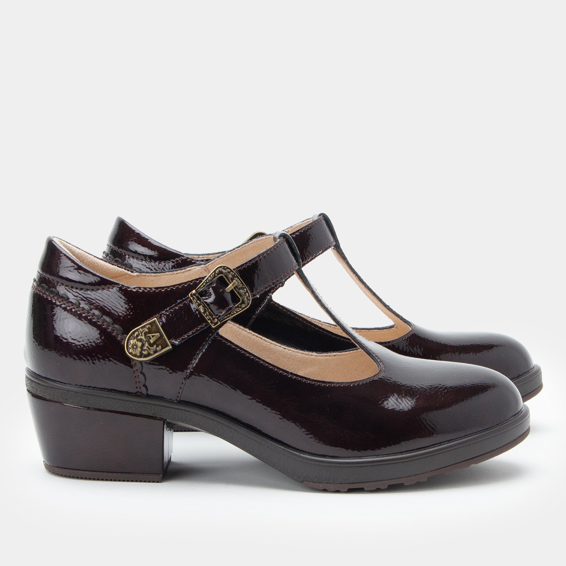 A pair of dark brown patent leather Mary Jane shoes with block heels and decorative buckle straps, shown against a light gray background.