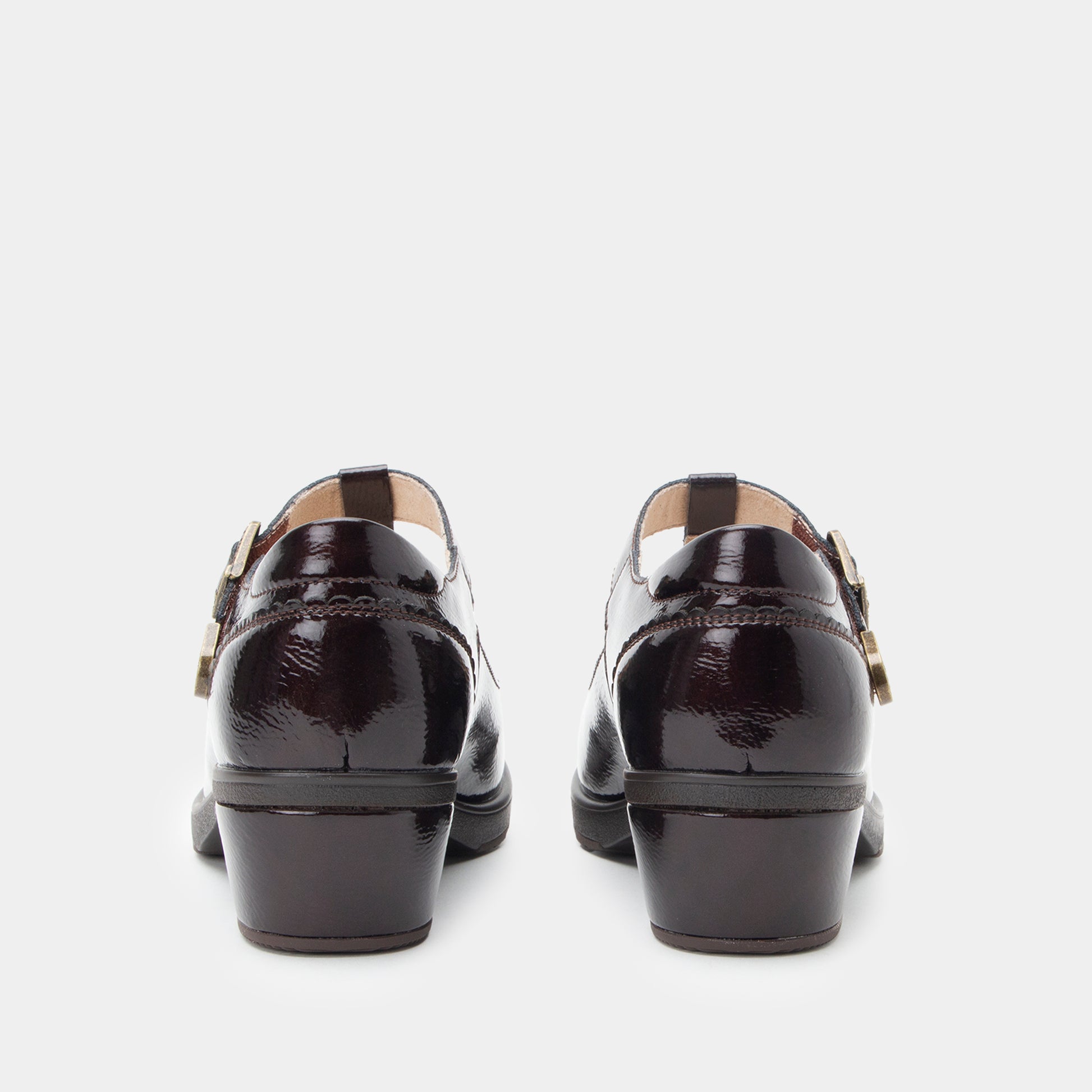 A pair of black patent leather Mary Jane shoes shown from the back, featuring low block heels and decorative gold-tone buckle straps, set against a plain light gray background.