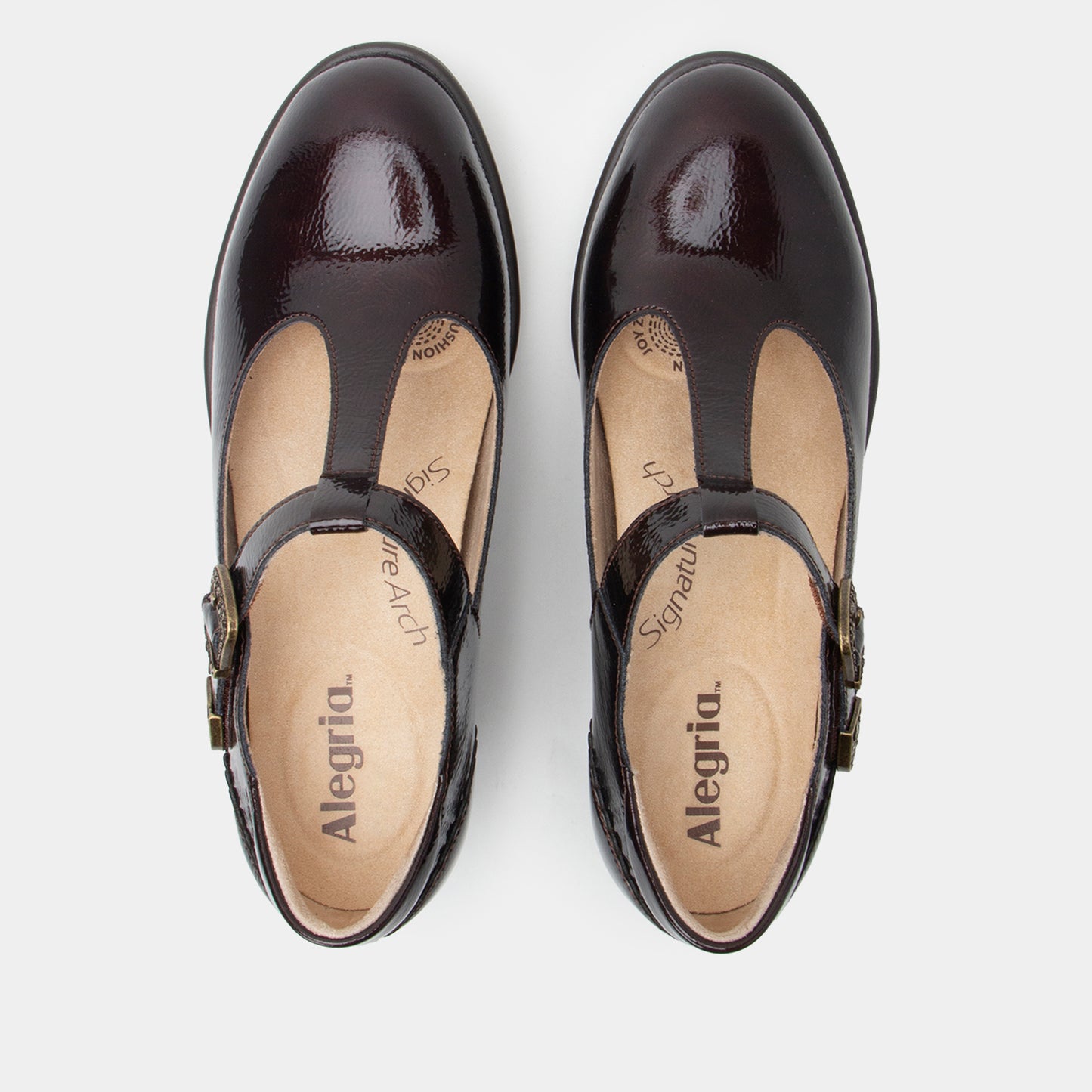 A pair of dark brown patent leather T-strap Mary Jane shoes with buckle straps, shown from a top-down view on a light gray background. The brand name 'Alegria' is visible on the beige insoles.