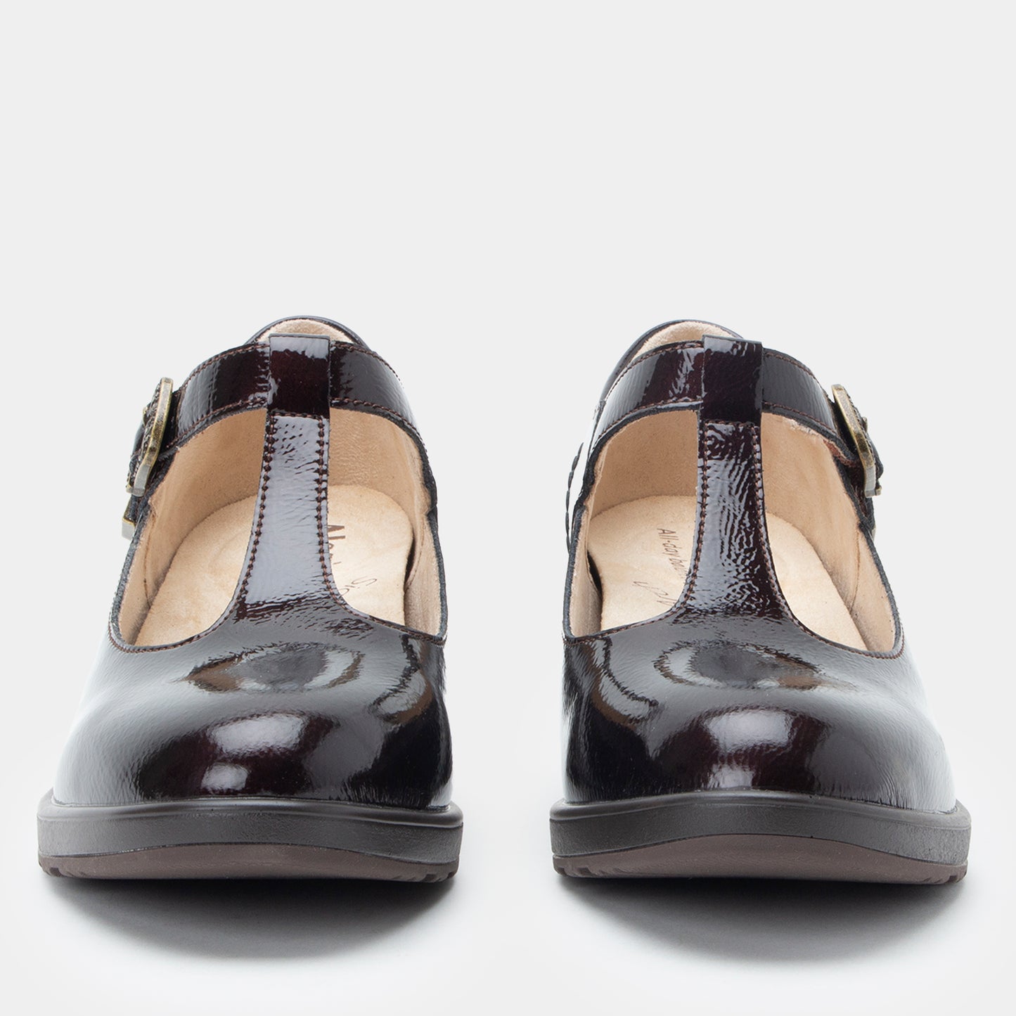 A pair of women's Mary Jane style shoes in glossy black patent leather, featuring a T-strap and a small gold buckle, shown from a front-facing perspective on a light gray background.
